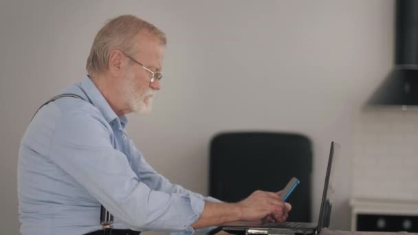 Modern old man handsome pensioner confident gadget user makes online purchases through Internet using a smartphone and laptop — Stock Video