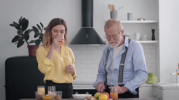 Homem idoso bonito com uma barba em óculos para a visão se diverte com a neta enquanto prepara o almoço de produtos saudáveis, bebendo suco e conversando na cozinha à mesa — Vídeo de Stock