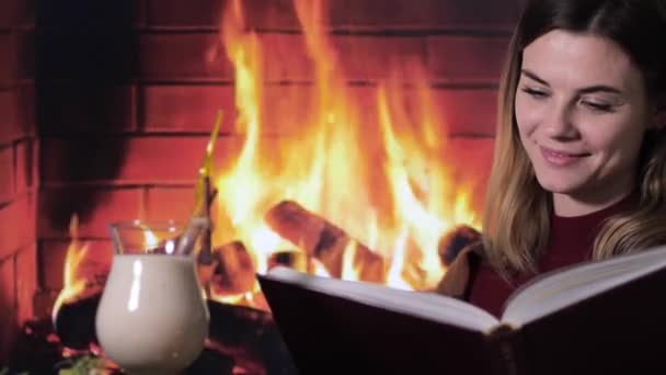 美しい若い女の子は暖炉の火に対して伝統的なカクテルの卵の近くの本でクリスマスの物語を読みます — ストック動画