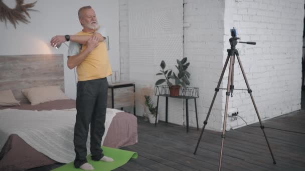 Влиятельные блоггер, пожилой мужчина показывает упражнение для суставов на телефоне камеры записи спортивного видеоблога для социальных сетей и последователей в своей комнате — стоковое видео