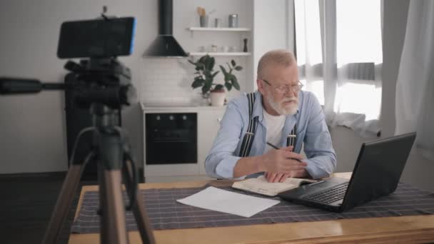 現代の年金ブロガーインフルエンサーは、ラップトップコンピュータで動作し、彼のフォロワーに本棚を説明する携帯電話にvlogを書きます — ストック動画