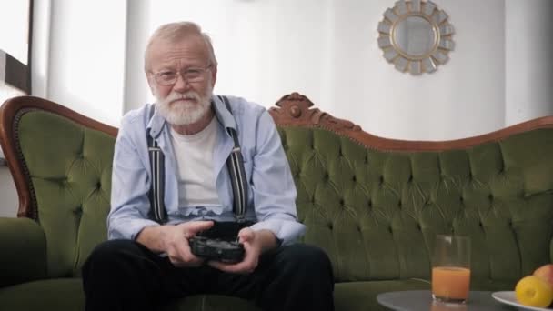 Atraktivní starý muž v brýlích pro vidění s vousy a joystick v ruce je baví hrát videohry, zatímco sedí na pohovce v místnosti — Stock video