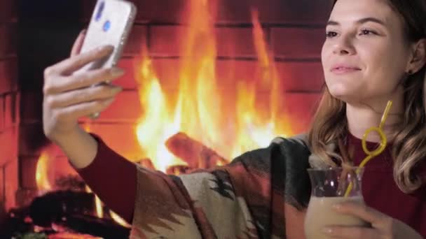 Jovem sorridente fêmea leva selfie no smartphone com tradicional eggnog cocktail na mão na lareira fundo — Vídeo de Stock