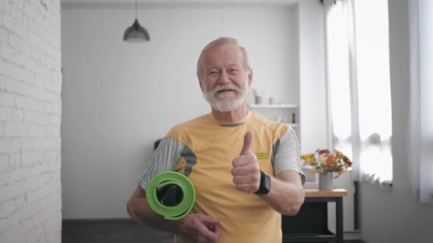 Gesundheitspflege, gut aussehender lächelnder alter Mann mit Yogamatte in den Händen und einer intelligenten Uhr nach dem Sport, um die Gesundheit zu erhalten und Daumenhoch-Geste, während er wieder drinnen steht — Stockvideo