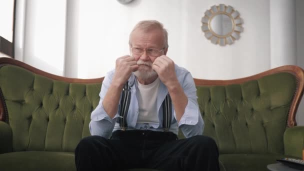 Deprese, starý důchodce s vousy a brýlemi pro vidění trpí a je smutný, protože život potíže sedí v místnosti na gauči — Stock video
