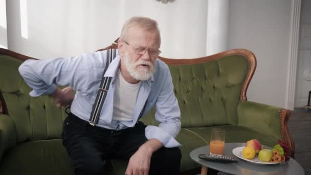 Grandfather in glasses and with beard is having pain in back grabs lower back with his hands while sitting on sofa in room — Stock Video
