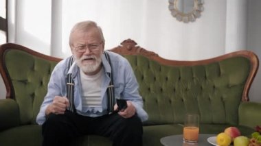 joyful old man, an elderly male with a beard in glasses for sight is watching a sports match on TV emotionally gesturing with hands while sitting on sofa, victory