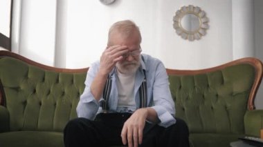 poor health, an old man suffering from headache migraine upset by life sitting on sofa in room, concept of loneliness
