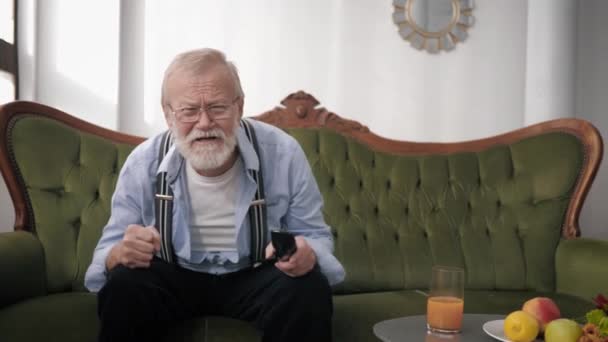 Joyful old man, an elderly male with a beard in glasses for sight is watching a sports match on TV emotionally gesturing with hands while sitting on sofa, victory — Stockvideo