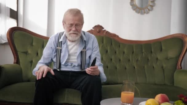 Old fan, grandfather with glasses for sight and beard with remote control very emotionally watches sports and gestures with hands while sitting on sofa against white wall — Stockvideo