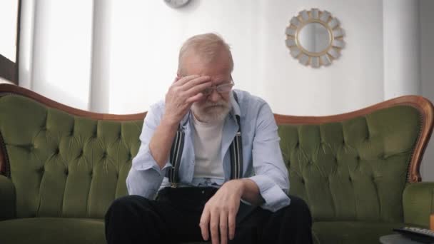 Poor health, an old man suffering from headache migraine upset by life sitting on sofa in room, concept of loneliness — Stock Video