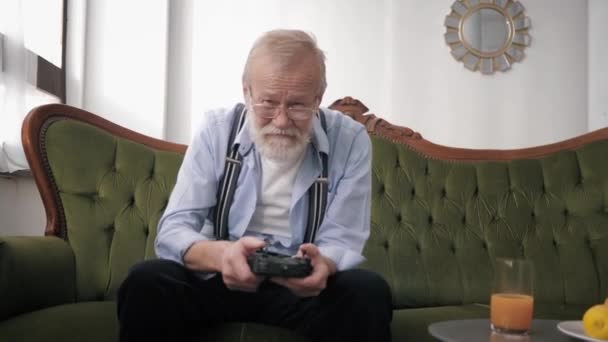 Χαμογελαστός γέρος gamer σε γυαλιά για όραση και γενειάδα, λάτρης των βιντεοπαιχνιδιών και παίζει ενεργά ενώ κάθεται στον καναπέ με χειριστήριο στα χέρια — Αρχείο Βίντεο
