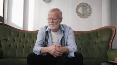 lonely elderly man with beard and glasses for vision suffers from depression and migraine with hands in head while sitting on sofa in room