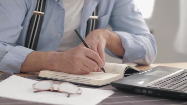 テーブルに座りながらパソコンで作業していると男性の手が閉まりノートをノートに書きます — ストック動画
