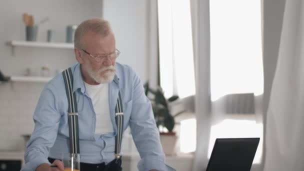 Moderno pensionista de edad avanzada con gafas para la visión trabaja duro en el ordenador portátil bebe jugo y escribe datos a cuaderno entiende por facturas — Vídeos de Stock