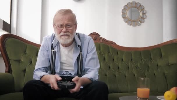 Modern grandfather gamer with beard and glasses for vision plays video games sitting on couch and holding joystick in hands — Stockvideo