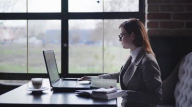 modern businesswoman provides advice online using the latest technology holds smartphone in her hands checking reports in mobile accounting working at laptop sitting at table by window