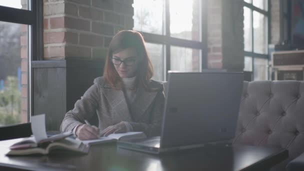 Fernstudium: Erfolgreiche Geschäftsfrau mit Brille macht sich online Notizen im Notizbuch, während sie im Café am Laptop arbeitet — Stockvideo