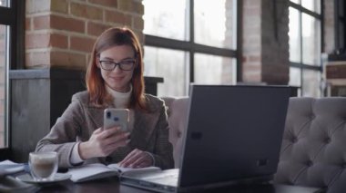 small business owner modern woman entrepreneur with glasses for vision working online behind laptop checking reports on smartphone sitting in cafe wifi