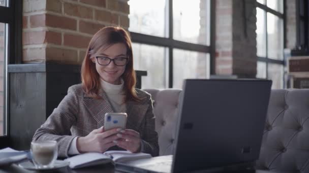 Online-Geschäft, junger vielversprechender Unternehmer arbeitet aus der Ferne mit Buchhaltungsunterlagen, die Berichte auf dem Smartphone prüfen, während er am Laptop im Café Wifi sitzt — Stockvideo