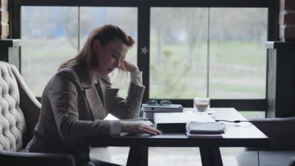 Joven chica triste está sentado molesto debido a la picadura de bloque de trabajo y problemas de contabilidad en los negocios, sostiene la cabeza con la mano mientras escribe en un cuaderno mientras está sentado en una mesa en un café — Vídeo de stock