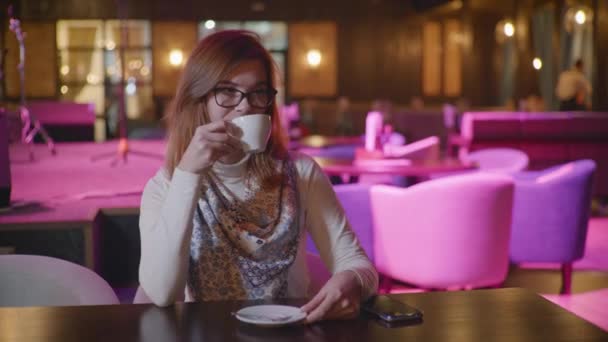 Attractive young woman with glasses enjoys coffee break relaxing in restaurant drinks coffee looks at camera and shows hand class then waves waving while sitting in a fashion institution — Stok video