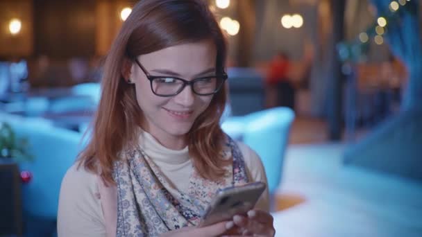 Gözlüklü gülümseyen kadın, sosyal ağdaki takipçilerle mesajlaşıyor. Akıllı telefondan gülümsüyor, el sallıyor ve güzel bir restoranda dinleniyor. — Stok video