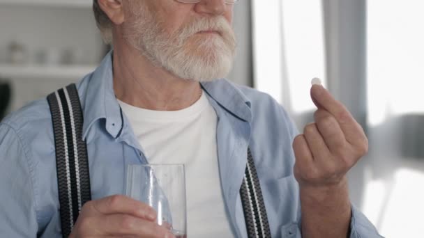 Gri sakallı yaşlı adam, sağlık için bardaktan su içiyor ve hapları alıyor. Beyaz odaya yakın, ağır çekimde. — Stok video