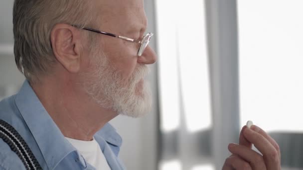 Health care, handsome mature man with gray beard close up takes a pill and drinks water from glass in kitchen — Stock Video