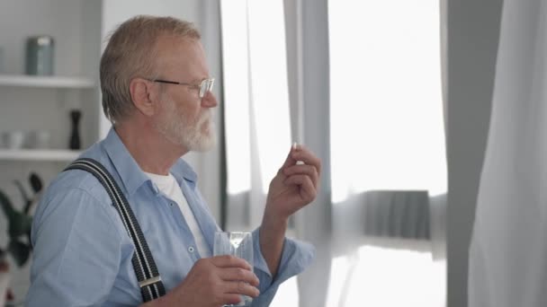 Homme mature avec barbe grise retraite prend une pilule avec de l'eau pour la santé et se sent mieux à la maison près de la fenêtre — Video