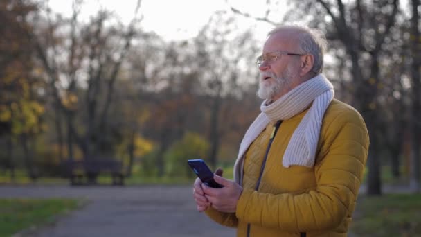 Nonno moderno con la barba grigia con un telefono cellulare in mano comunica tramite videochiamata mentre cammina nel parco autunnale — Video Stock