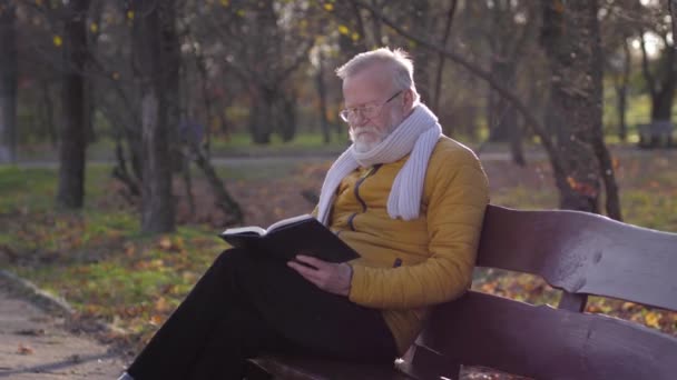 Stáří, důchodce muž v brýlích s poznámkovým blokem a tužkou v ruce něco plánuje, zatímco sedí na lavičce v podzimním parku — Stock video