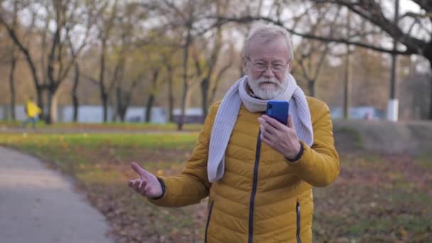 Γέρος χρησιμοποιεί το κινητό τηλέφωνο για μια βιντεοκλήση, αρσενικό με μια γκρίζα γενειάδα περπάτημα στο πάρκο δείχνει την ομορφιά της φύσης — Αρχείο Βίντεο