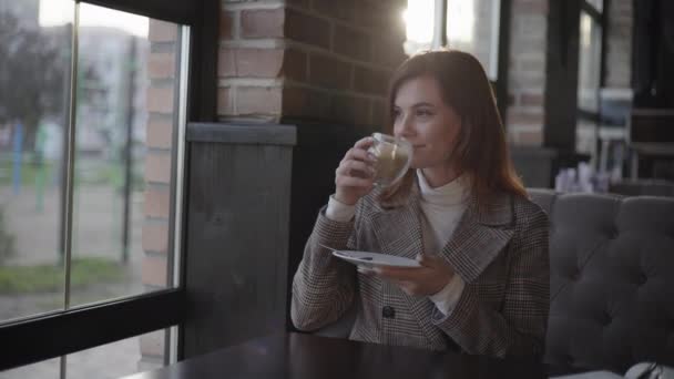 Hermosa mujer bebe café, chica con una taza de capuchino en las manos sonríe soñadoramente y mira por la ventana disfrutando de relajarse sentado en la mesa en la acogedora cafetería — Vídeos de Stock