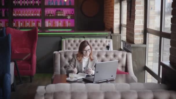 Exitosa mujer de negocios en vasos bebe café en el restaurante trabajando en un ordenador portátil y teléfono inteligente, disfruta de trabajar en línea sentado en la mesa por la ventana, el trabajo moderno — Vídeo de stock
