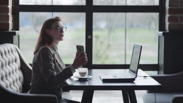 Привлекательная девушка весело разговаривает на видео-звонок с ее другом онлайн через ноутбук показывает смартфон, сидя за столом в уютном кафе — стоковое видео