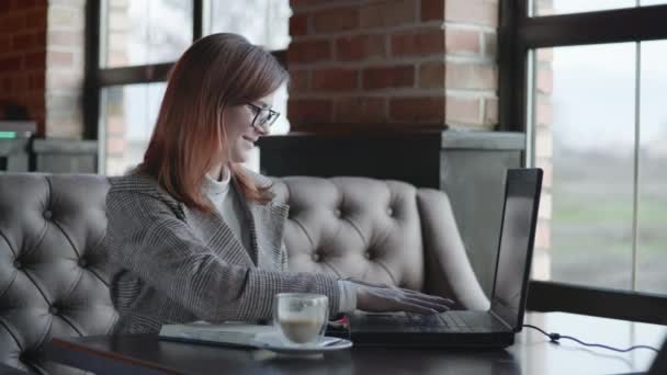 Uzaktan çalışma, görüş için gözlüklü başarılı kız bir dizüstü bilgisayar kullanarak online işten zevk alıyor ve masada otururken gülümseyen ve çerçeveye bakan bir deftere veri yazıyor — Stok video