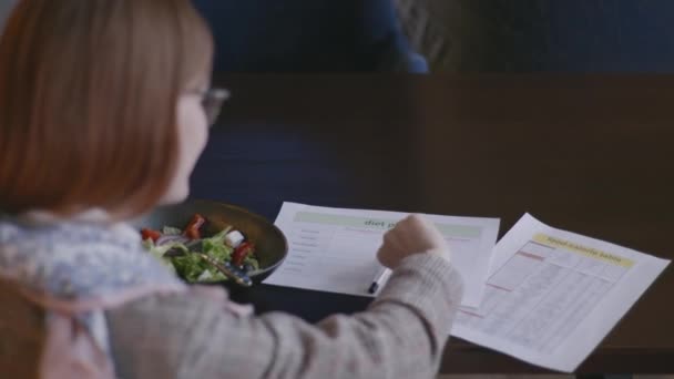 Comida saudável, menina bonita jovem senta-se na dieta e verifica o conteúdo calórico de produtos em salada vegetariana de legumes frescos e escreve o resultado em plano de dieta enquanto sentado à mesa em aconchegante — Vídeo de Stock