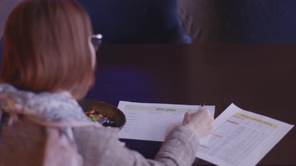 Gewichtsverlies, een aantrekkelijke jonge vrouw neemt goede voeding en merkt calorie inhoud van producten in vegetarische salade van verse groenten opschrijven hoeveelheid voedsel gegeten in dieet plan, terwijl — Stockvideo