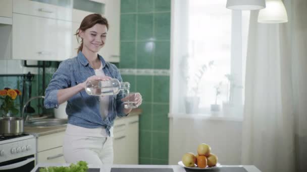 Jovem mulher bonito derrama água em vidro, em seguida, bebe olha para a câmera e sorri enquanto está na cozinha à mesa, estilo de vida saudável — Vídeo de Stock