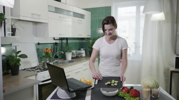 Menina sorridente atraente se diverte enquanto cozinha e assiste a um vídeo de receita de um blog popular sobre alimentação saudável, mostra com a mão e gosta de estar em uma mesa na cozinha — Vídeo de Stock