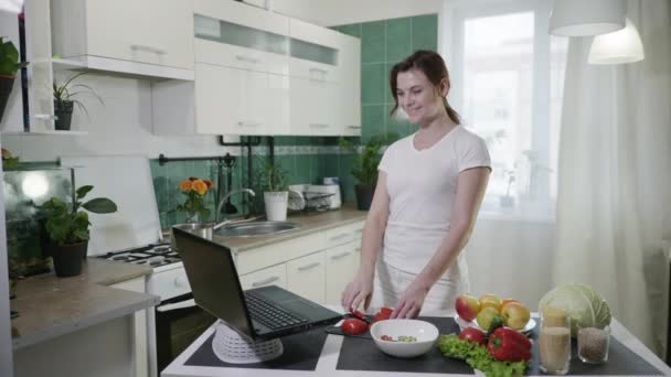 Freudige Hausfrau hat Spaß bei der Zubereitung eines gesunden Frühstücks aus vegetarischen Produkten mit Rezept von vlog lächelnd und schneidet frisches Gemüse am Tisch in der Küche stehen — Stockvideo