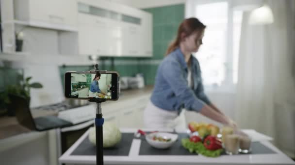 Attraktiv flicka skriver vlog video på smartphone för anhängare berättar om diet plan visar på kameran under direktsändning vid bordet med användbara produkter i köket — Stockvideo