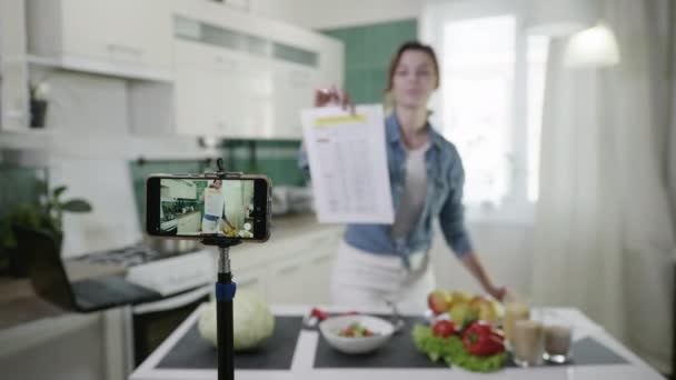 现代家庭主妇博客在智能手机上记录视频内容，以讨论适当的营养，这显示了一个饮食计划在相机中出现，然后竖起大拇指站在桌子旁 — 图库视频影像
