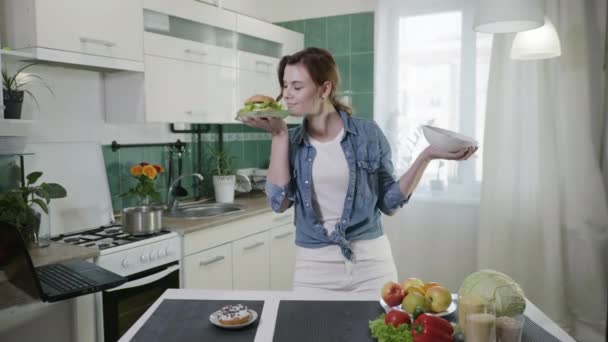 Ein schönes Mädchen wählt fröhlich zwischen einem Burger und einem vegetarischen Salat mit frischem Gemüse, hält sie in den Händen und lacht neugierig ins Gesicht — Stockvideo