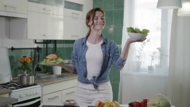 Sağlık hizmetleri, çekici bir kız doğru beslenmeyi izler ve elinde taze sebzelerden oluşan vejetaryen salatası tutar ve bir hamburger seçimini yapar. — Stok video