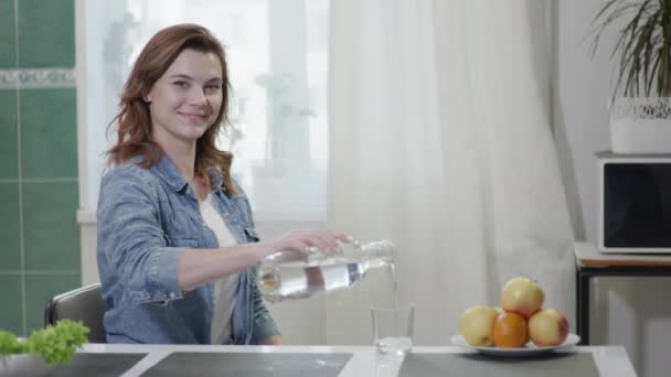 Aantrekkelijk lachend meisje giet schoon water in een glas, drinkt en kijkt naar de camera terwijl zitten aan tafel in de keuken, gezondheid concept — Stockvideo