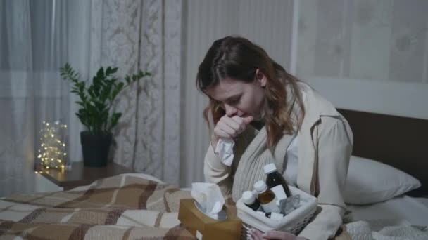 Sick girl suffers from flu or illness, measures her temperature with thermometer with medicines in hands, sneezes and quenches while sitting on a sofa in room — Stock Video