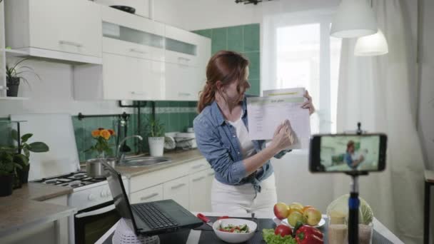 Blog de mujeres, joven ama de casa atractiva mantiene blog y habla sobre los planes de dieta y calorías mientras está de pie en la cocina en la mesa con verduras frescas, personas y redes sociales — Vídeo de stock