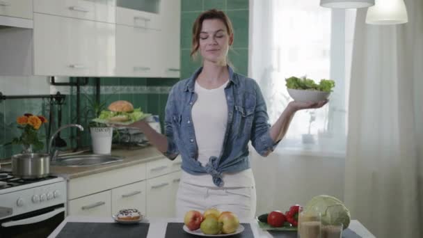 Food products, lovely young girl holding hamburger and salad of fresh vegetables is trying to make choice between products, is looking at camera and smiling while standing at table in kitchen — Stock Video
